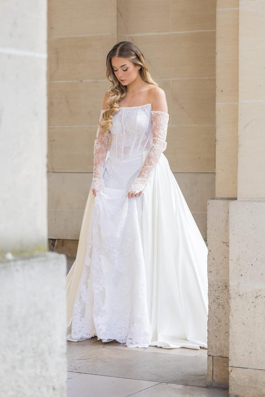 Mariée en robe de dentelle intégrale, regard baissé devant un mur de pierre